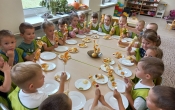 Czterolatki w zielonych fartuszkach siedza wokol  stolu i prezzentuja wykonane samodzielnie szaszlyki owocowe.